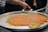 A large metal tray contains a traditional dessert, possibly kunafa, with a golden-orange top layer and sprinkled with green pistachio bits. A person is cutting and serving a piece onto a plate, while stacks of empty plates are visible in the background.