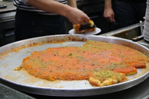 A large metal tray contains a traditional dessert, possibly kunafa, with a golden-orange top layer and sprinkled with green pistachio bits. A person is cutting and serving a piece onto a plate, while stacks of empty plates are visible in the background.
