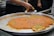 A large metal tray contains a traditional dessert, possibly kunafa, with a golden-orange top layer and sprinkled with green pistachio bits. A person is cutting and serving a piece onto a plate, while stacks of empty plates are visible in the background.