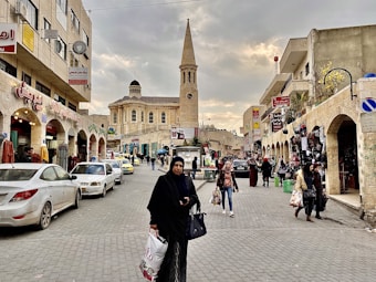 A bustling urban street scene with a mixture of traditional and modern architecture. The central focus is a tall beige stone building with a clock tower, resembling a large religious structure. Surrounding this are two-story buildings with shops on the ground floor, displaying signs in Arabic. People of various ages walk along the cobbled street, some carrying shopping bags. Several parked and moving cars are visible. The sky is overcast with a hint of evening light.