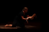 A person is kneeling on a dimly lit stage, holding a white cloth in their hands. The focus is on the expressive posture as they appear to be performing a dramatic scene.
