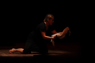 A person is kneeling on a dimly lit stage, holding a white cloth in their hands. The focus is on the expressive posture as they appear to be performing a dramatic scene.