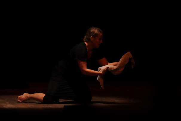 A person is kneeling on a dimly lit stage, holding a white cloth in their hands. The focus is on the expressive posture as they appear to be performing a dramatic scene.
