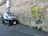 A small electric car is parked next to a wall with graffiti depicting a cartoon character with yellow skin and spiky hair, holding a can of spray paint. The charger cable is connected to the car from a nearby electrical box.