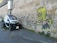 A small electric car is parked next to a wall with graffiti depicting a cartoon character with yellow skin and spiky hair, holding a can of spray paint. The charger cable is connected to the car from a nearby electrical box.