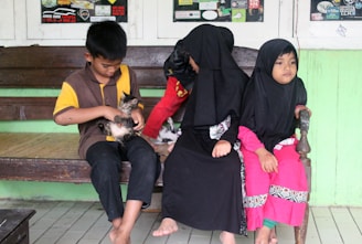 Three children are sitting on a wooden bench. Two of them are wearing black headscarves, and brightly colored clothing, while the boy is holding a small kitten. The background shows a green painted wall and posters on the windows.