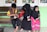 Three children are sitting on a wooden bench. Two of them are wearing black headscarves, and brightly colored clothing, while the boy is holding a small kitten. The background shows a green painted wall and posters on the windows.