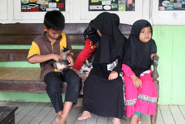 Three children are sitting on a wooden bench. Two of them are wearing black headscarves, and brightly colored clothing, while the boy is holding a small kitten. The background shows a green painted wall and posters on the windows.