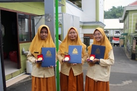 Three young women wearing matching mustard-colored hijabs and uniforms stand outside. Each holds a dark blue certificate folder embossed with a crest and a round medal in a red case. The background includes a building with green walls and an Indonesian flag.