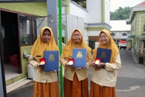 Three young women wearing matching mustard-colored hijabs and uniforms stand outside. Each holds a dark blue certificate folder embossed with a crest and a round medal in a red case. The background includes a building with green walls and an Indonesian flag.