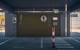 A covered parking area features a large, numbered green door with a circular sign displaying the number '1'. To the left of the door, there's a wall-mounted device, possibly an intercom or control unit. Above the door, an emergency exit sign is visible. A striped bollard stands in the foreground, painted red and white, and pavement markings are visible on the ground.