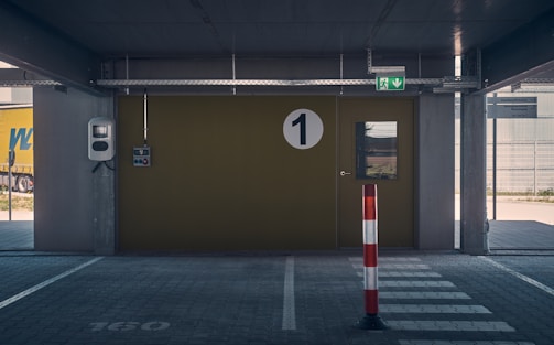 A covered parking area features a large, numbered green door with a circular sign displaying the number '1'. To the left of the door, there's a wall-mounted device, possibly an intercom or control unit. Above the door, an emergency exit sign is visible. A striped bollard stands in the foreground, painted red and white, and pavement markings are visible on the ground.