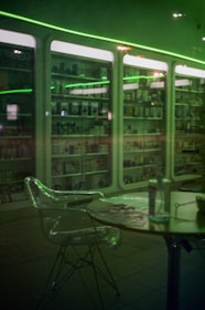A dimly lit interior of a store or showroom with glass display cabinets filled with various products. The scene is cast in a green hue due to the overhead neon lights. In the foreground, there is a transparent chair and a table with items on it, including a bottle.