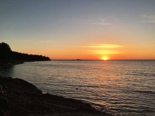 The image depicts a serene sunset over a calm sea, with the sun positioned near the horizon. The sky is a blend of soft blue and warm orange hues, creating a tranquil atmosphere. Dark silhouettes of trees and the coastline are visible on the left side, while gentle waves ripple across the water's surface.