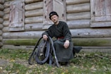 A man in traditional clothing is sitting on the ground against a wooden log cabin, holding a set of bagpipes. The background features rustic wooden walls with closed shutters on small windows. Fallen leaves are scattered on the ground, suggesting an autumn setting.