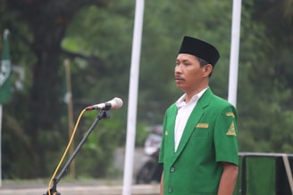 A man stands in front of a microphone outdoors, wearing a green jacket with an embroidered logo and a name tag. His expression is serious and focused.