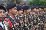 A group of people dressed in camouflage uniforms stand in a formation. They are wearing black berets and have badges on their uniforms. The environment is outdoors, with greenery and stone wall in the background.