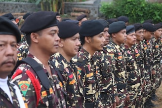A group of people dressed in camouflage uniforms stand in a formation. They are wearing black berets and have badges on their uniforms. The environment is outdoors, with greenery and stone wall in the background.