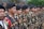 A group of people dressed in camouflage uniforms stand in a formation. They are wearing black berets and have badges on their uniforms. The environment is outdoors, with greenery and stone wall in the background.