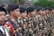 A group of people dressed in camouflage uniforms stand in a formation. They are wearing black berets and have badges on their uniforms. The environment is outdoors, with greenery and stone wall in the background.