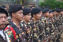 A group of people dressed in camouflage uniforms stand in a formation. They are wearing black berets and have badges on their uniforms. The environment is outdoors, with greenery and stone wall in the background.