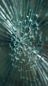 A close-up view of shattered glass, with multiple cracks radiating from a central point, creating a pattern that looks intricate and chaotic.