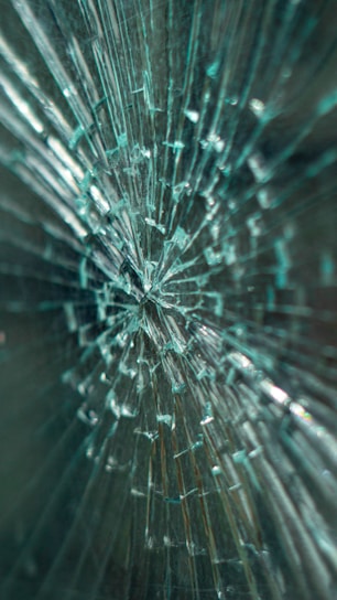 A close-up view of shattered glass, with multiple cracks radiating from a central point, creating a pattern that looks intricate and chaotic.