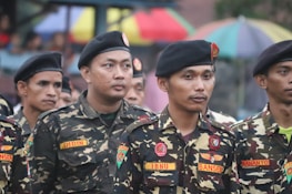 A group of men wearing military-style uniforms with camouflage patterns and black berets standing together in a formation. The uniforms have patches and name tags visible. In the background, there are colorful umbrellas.