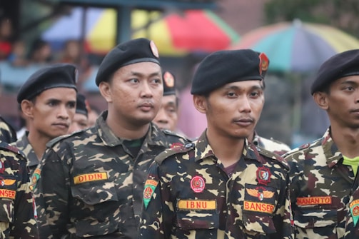A group of men wearing military-style uniforms with camouflage patterns and black berets standing together in a formation. The uniforms have patches and name tags visible. In the background, there are colorful umbrellas.