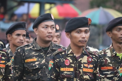 A group of men wearing military-style uniforms with camouflage patterns and black berets standing together in a formation. The uniforms have patches and name tags visible. In the background, there are colorful umbrellas.