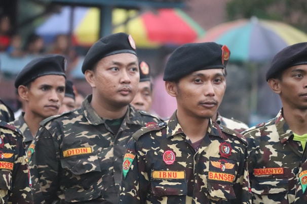 A group of men wearing military-style uniforms with camouflage patterns and black berets standing together in a formation. The uniforms have patches and name tags visible. In the background, there are colorful umbrellas.
