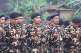Several men dressed in camouflage military uniforms and black berets stand outdoors with their hands raised, appearing to be in a moment of prayer or reflection. The background features lush greenery and a building with a thatched roof, suggesting a rural setting.