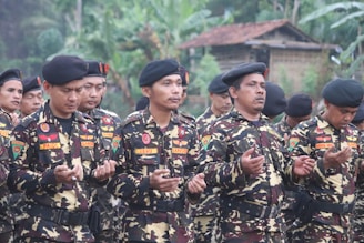 Several men dressed in camouflage military uniforms and black berets stand outdoors with their hands raised, appearing to be in a moment of prayer or reflection. The background features lush greenery and a building with a thatched roof, suggesting a rural setting.