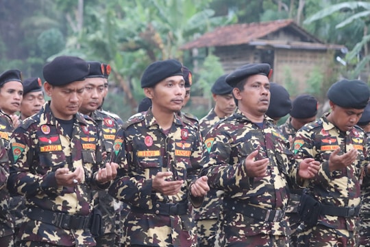 Several men dressed in camouflage military uniforms and black berets stand outdoors with their hands raised, appearing to be in a moment of prayer or reflection. The background features lush greenery and a building with a thatched roof, suggesting a rural setting.