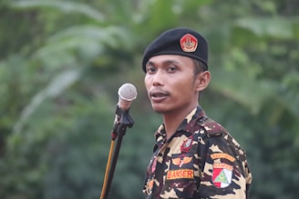 A person stands outdoors, wearing a camouflage uniform with various patches on the chest and sleeves. A black beret with an emblem is worn on the head. A microphone on a stand is positioned in front of the person. The background is a blur of lush green foliage.