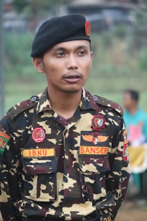 A person in a camouflage uniform is standing outdoors. The uniform has various patches, including one that reads 'IBNU' and another that says 'BANSER'. The individual is wearing a black beret with a red emblem.