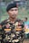 A person in a camouflage uniform is standing outdoors. The uniform has various patches, including one that reads 'IBNU' and another that says 'BANSER'. The individual is wearing a black beret with a red emblem.