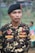 A person in a camouflage uniform is standing outdoors. The uniform has various patches, including one that reads 'IBNU' and another that says 'BANSER'. The individual is wearing a black beret with a red emblem.