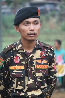 A person in a camouflage uniform is standing outdoors. The uniform has various patches, including one that reads 'IBNU' and another that says 'BANSER'. The individual is wearing a black beret with a red emblem.