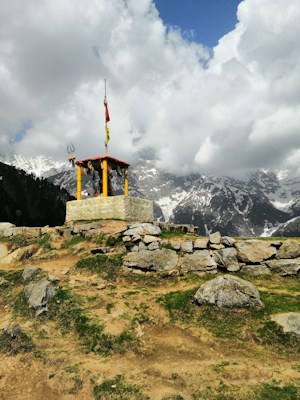 A small shrine or altar structure with a red roof and yellow columns is situated on a rocky hilltop. It features flags and tridents, typical of religious symbolism. The background consists of majestic snow-capped mountains partially obscured by large, fluffy clouds. The foreground contains rough terrain with scattered rocks and patches of grass.