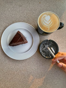 A piece of chocolate cake is placed on a white plate next to a cup of latte with heart-shaped latte art. Adjacent to the cup is an ashtray containing cigarette butts, with a hand holding a burning cigarette.