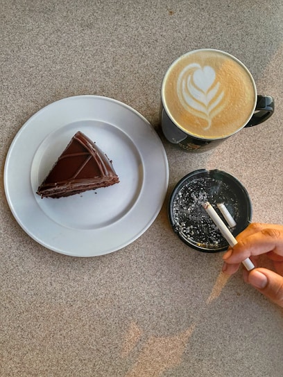 A piece of chocolate cake is placed on a white plate next to a cup of latte with heart-shaped latte art. Adjacent to the cup is an ashtray containing cigarette butts, with a hand holding a burning cigarette.