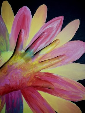 A hand covered in multicolored paint is laid on top of a large, vibrant painting of a flower. The petals of the flower are in shades of pink, yellow, and a touch of green against a dark background. The hand's paint colors blend harmoniously with the painted petals, creating an artistic and playful effect.