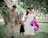 A family consisting of two adults and a child walks through a park. The woman wears a white dress and holds a large pink balloon shaped like the number five. The man wears a floral shirt and watches the child, who is in a black dress and holding hands with both adults. They are surrounded by greenery, including trees and grass, suggesting a peaceful environment.