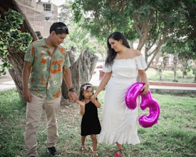 A family consisting of two adults and a child walks through a park. The woman wears a white dress and holds a large pink balloon shaped like the number five. The man wears a floral shirt and watches the child, who is in a black dress and holding hands with both adults. They are surrounded by greenery, including trees and grass, suggesting a peaceful environment.