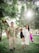 A happy family enjoying time in a lush green park. A man in a floral shirt and beige pants is holding hands with a little girl in a dark dress, and a woman in a white dress. They are playfully swinging the child. Sunlight filters through the foliage, creating a serene and warm atmosphere.