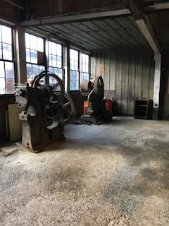 An old workshop with large, vintage machinery and wooden walls. The floor appears to be made of concrete, and there are large windows letting in natural light. The space looks worn and aged, suggesting it has been used for industrial purposes.