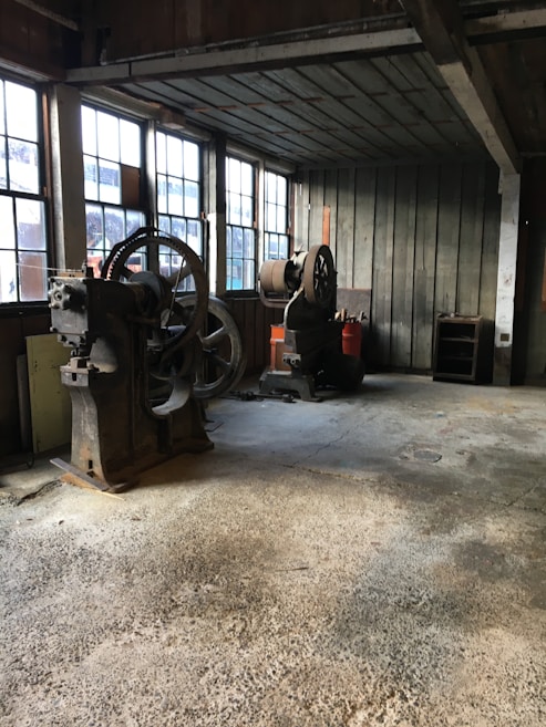An old workshop with large, vintage machinery and wooden walls. The floor appears to be made of concrete, and there are large windows letting in natural light. The space looks worn and aged, suggesting it has been used for industrial purposes.