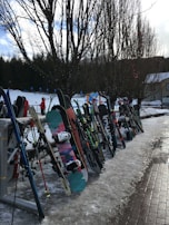 A variety of skis and snowboards are neatly lined up and resting against a rack outdoors, with snow covering the ground. Bare trees stand in the background, and a snowy landscape suggests a winter sport setting. A few people in warm clothing are visible in the distance, possibly near a ski resort area.