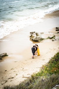 A couple stands on a sandy beach near the water's edge, with a photographer capturing their moments. The woman is wearing a bright yellow dress, while the man is dressed casually. The waves gently lap onto the shore, and there are some rocky formations and patches of vegetation in the foreground.